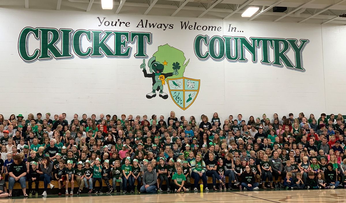 Hundreds of students sit in a gymnasium under the Fall Creek Crickets logo.