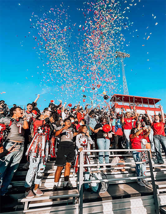 Photograph of students cheering in bleachres at a football game with confetti in the air above them