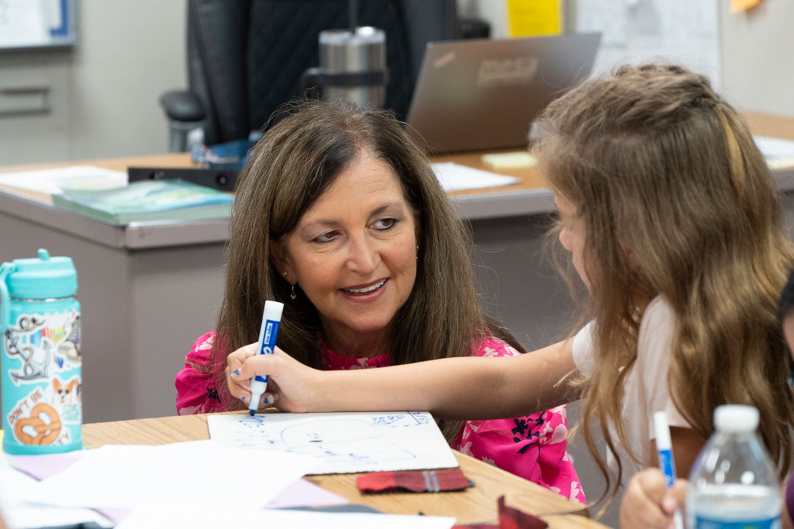 Photograph of Dr. Andi Fourlis kneeling next to a student at her desk 