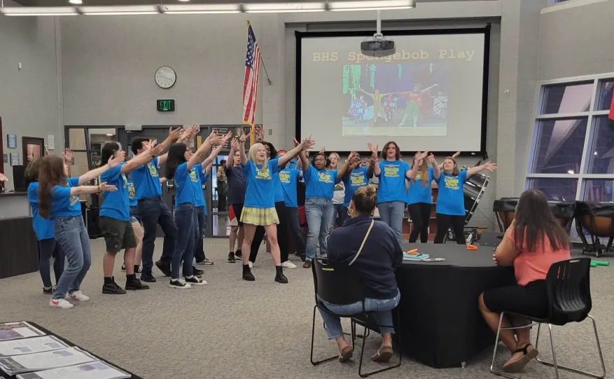 Several students in blue T-shirts rehearse a theatre performance for their teachers.